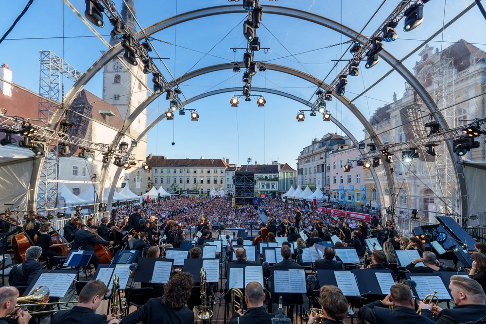 Aussicht von einer Open-Air-Bühne mit vielen Musikern darauf auf einen Platz mit sitzendem Publikum