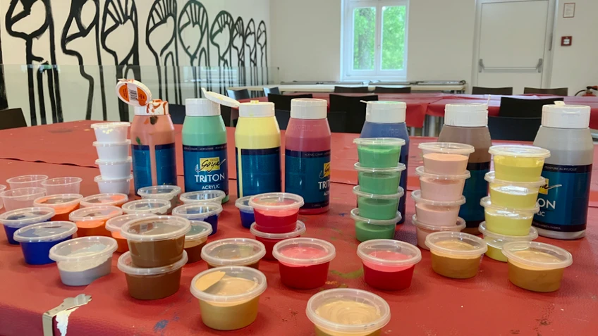 Tisch mit zahlreichen offenen Acrylfarben in Flaschen und kleinen Bechern, sortiert in verschiedenen bunten Farbtönen in einem Atelierraum.