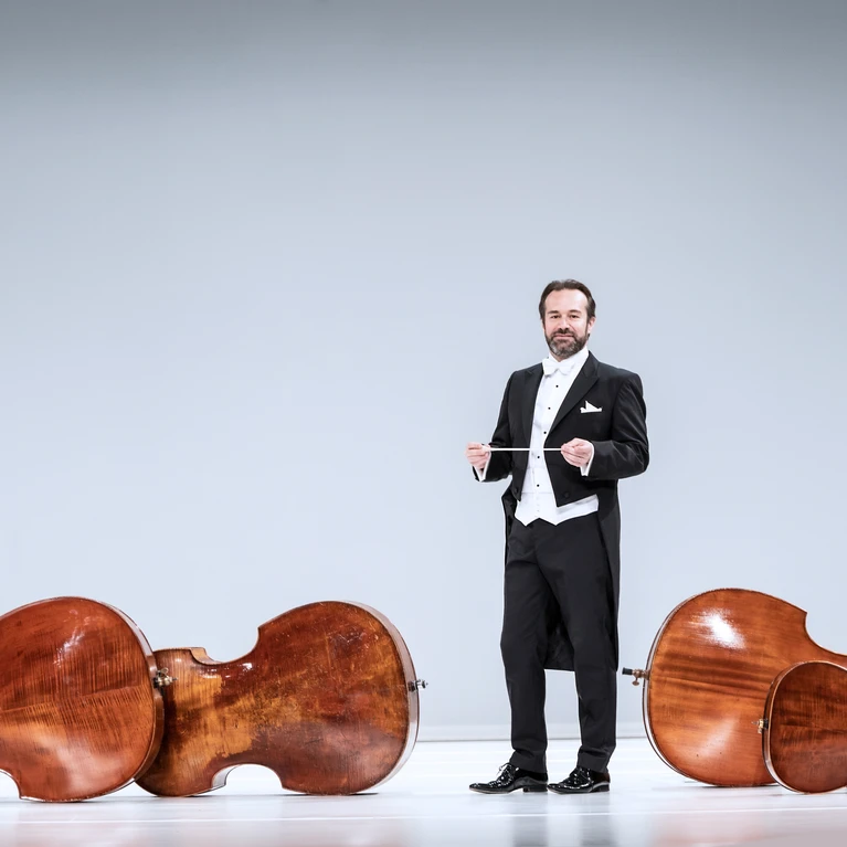 Fabien Gabel in a tailcoat in front of a white background, surrounded by orchestral instruments on the floor. 