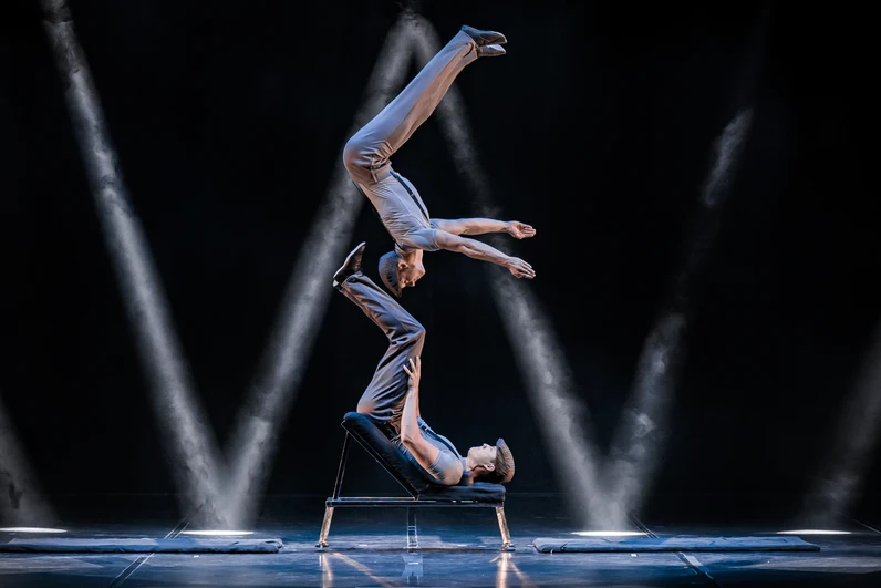 Zwei Artisten in grauer Kleidung führen auf einer Bühne eine akrobatische Partnerbalance vor, bei der der eine auf dem Rücken liegend den anderen mit den Füßen senkrecht nach oben in der Luft hält, während Spotlights die Szene dramatisch ausleuchten.