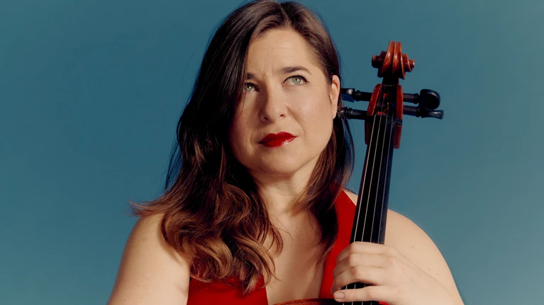 Photo of Alisa Weilerstein, soloist with the violoncello, with her instrument in front of a light blue background.