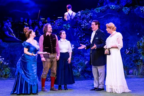 A group scene on stage featuring several people in different costumes, engaged in dialogue with one another. The stage is decorated with green elements and bathed in blue light. 