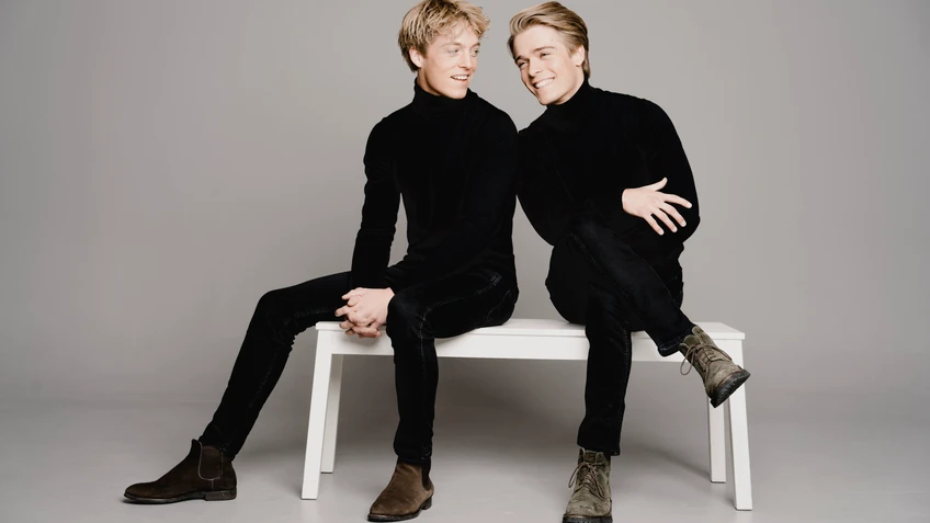 Photograph of brothers Lucas and Arthur Jussen, virtuoso pianists, seated on a white bench in front of a light grey wall.