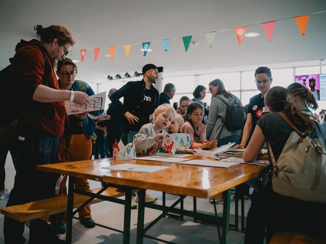 Photo from the BIG BANG Festival, showing a table surrounded by children and adults, with a colourful garland hanging in the background.
