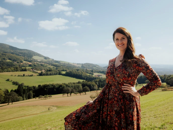 Dunkelhaarige Dame in geblümtem Kleid, dahinter grünes Hügelland