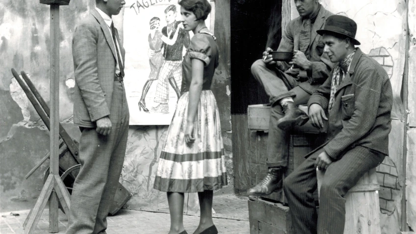 Ausschnitt aus einem alten schwarz-weiß-Film, drei Männer, eine Frau vor einer Türe, zwei Männer sitzen auf Holzkisten, eine Theaterlaterne daneben