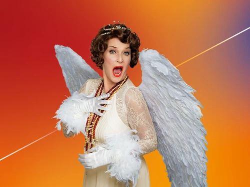 ine Person in einem weißen Engelskostüm mit großen Federflügeln und Krone posiert ausdrucksstark vor einem farbintensiven Hintergrund.