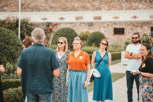 Kulturvermittler mit Rücken zu Betrachter erzählt einer Gruppe von 7 Personen etwas im Garten