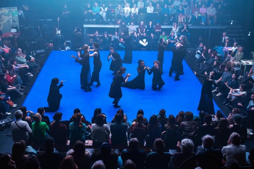 Bühnenfoto von "Chapters of Celebration", choreografiert von Benjamin Abel Meirhaege. Tänzer:innen, schwarz gekleidet, auf einer auffällig blauen Bühne. Zu sehen ist außerdem das diese Bühne an allen Seiten umringende Publikum 