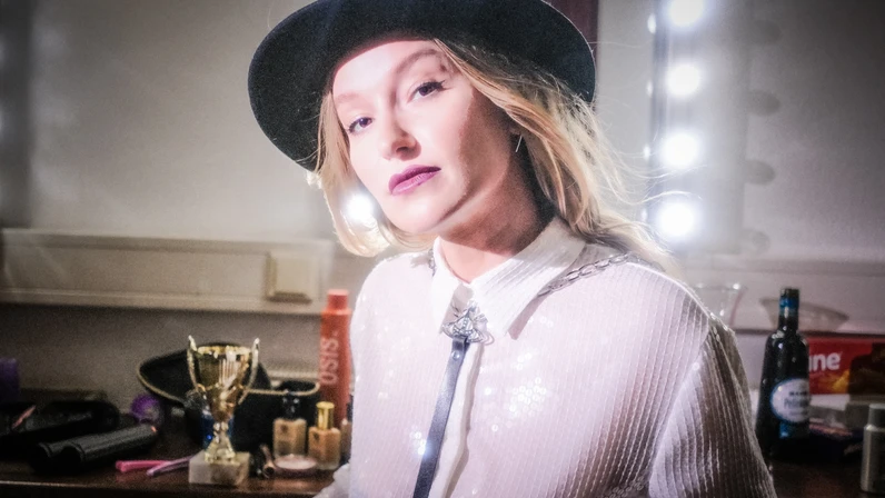 Malarina sitzt mit langem blondem Haar und schwarzem Hut vor einem beleuchteten Schminktisch und blickt selbstbewusst in die Kamera, umgeben von Make-up‑Utensilien, einer Trophäe und Getränken.