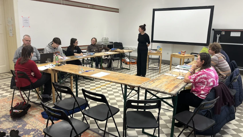 Schreib-Werkstatt vom Verein Ohrenschmaus wo eine Gruppe von Personen in einem Seminarraum an zu einem U-förmigen Kreis angeordneten Holztischen sitzt. Eine stehende Vortragende spricht zur Gruppe. Auf den Tischen liegen Schreibmaterialien