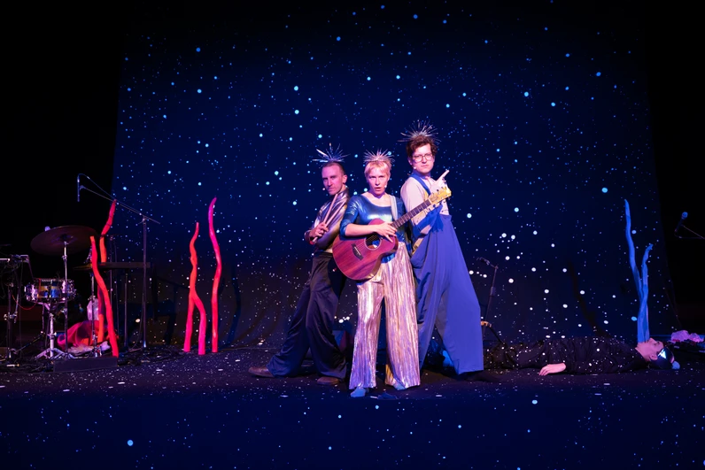 Drei als fantasievolle Weltraumwesen verkleidete Musikerinnen und Musiker stehen auf einer dunkelblauen Bühne voller glitzernder Sterne, während zwei Gitarren gespielt werden und im Hintergrund ein weiterer Darsteller am Boden liegt.