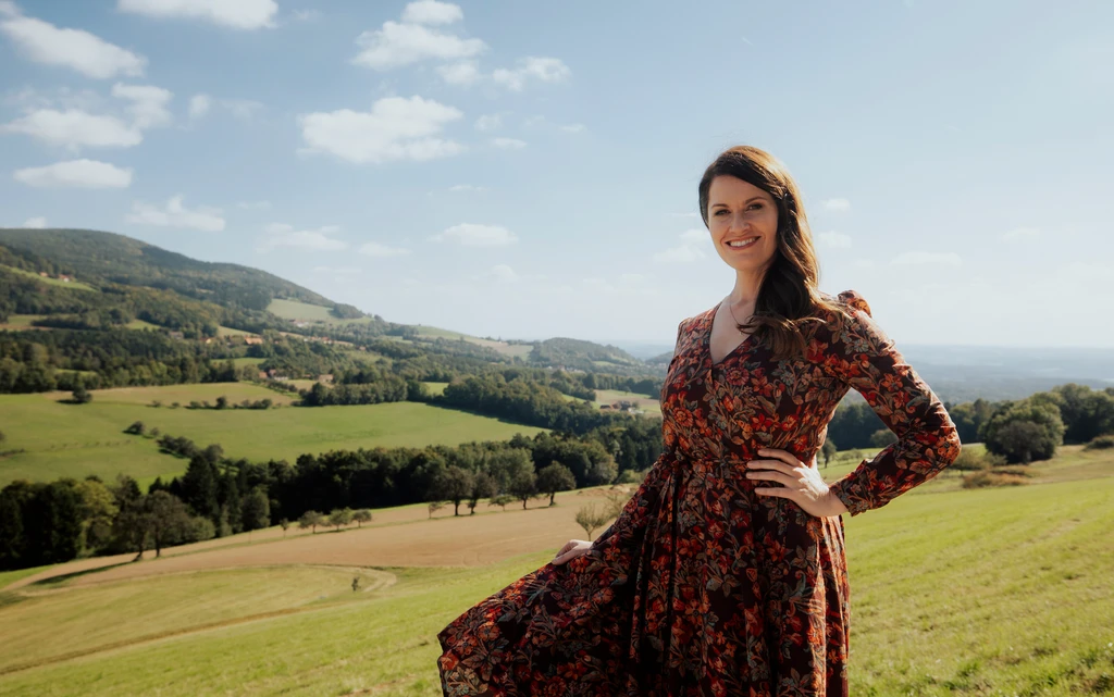 Frau in geblümtem Kleid mit dunklen langen Haaren auf einer Wiese, im Hintergrund Hügelland