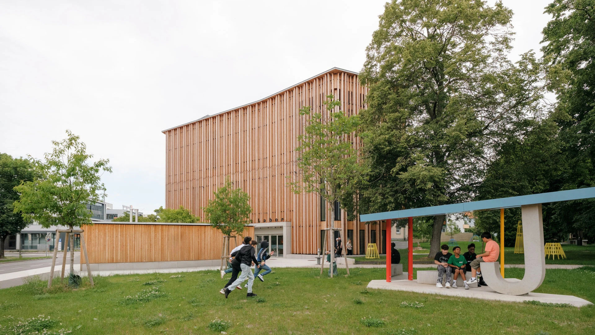 Modernes Gebäude mit vertikaler Holzfassade inmitten einer Grünfläche, Kinder spielen und sitzen im Vordergrund.