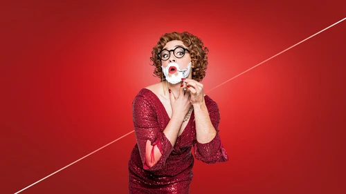 Eine Person in rotem Glitzerkleid mit Perlenkette und Locken rasiert sich das Gesicht vor rotem Hintergrund.