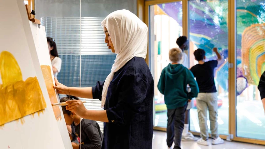 In the foreground, a girl paints at an easel, while in the background, three boys look through a glass door at a colourful textile sculpture by Toshiko Horiuchi MacAdam.