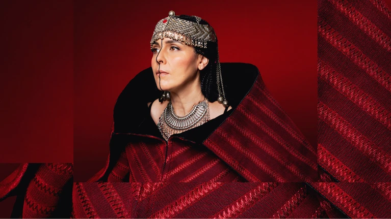 A portrait photograph of the Algerian musician Souad Massi wearing a striking traditional red robe, a necklace and headdress, and light face paint, against a red background; she is turned slightly to the left and looking past the camera.