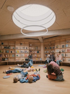 Kinder liegen am Boden und schauen zum runden Dachfenster der KinderKunstLabor-Bibliothek hinaus. Im Hintergrund sind Wände voller Bücher.