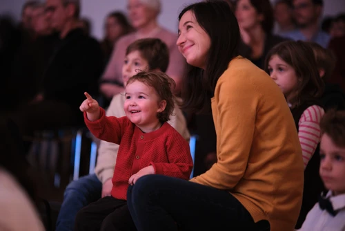 Publikumsfoto vom "Tonspiele"-Projekt "Schnee" - einem Projekt des Musikvermittlungsprogramm des Tonkünstler-Orchester Niederösterreich. Zu sehen sind fröhliche Kinder, die gespannt zuhören und -schauen. Ein Kind sitzt auf dem Schoß seiner Mutter und zeigt in Richtung der Bühne.