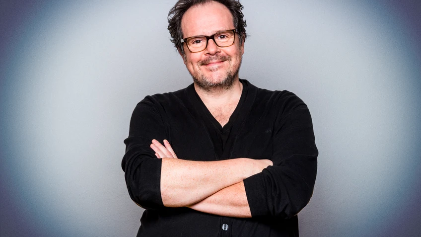 Thomas Mraz steht lächelnd mit Brille und dunkler Kleidung vor einem neutralen grauen Hintergrund und hat die Arme locker vor der Brust verschränkt.