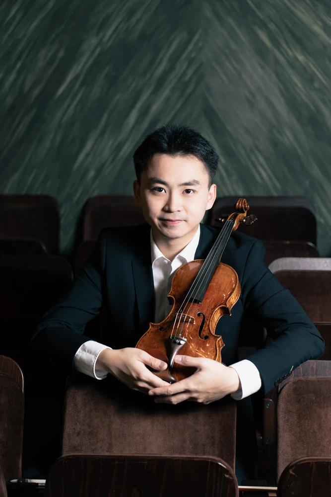 Mann mit Anzug sitzt mit einer Violine zwischen Stühlen