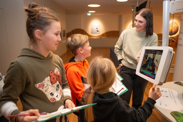 Drei Kinder haben ein Klemmbrett und nehmen an der Rätselrallye im Haus der Wildnis teil. Ein Guide erklärt ihnen die Inhalte mit einem in der Ausstellung angebrachten, interaktiven Tablet. 