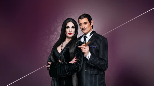Two people pose as Morticia and Gomez Addams in classic costumes against a dark purple floral background.