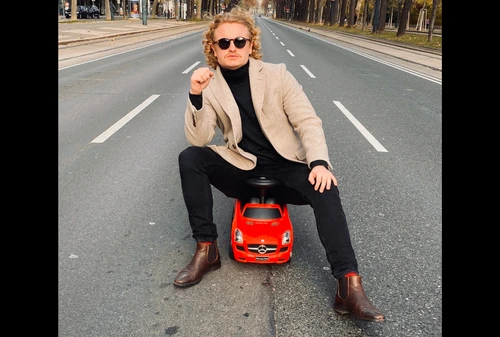Mann mit blonden Locken und dunkler Sonnenbrille in schicker Kleidung sitzt auf einem Modellauto am Wiener Ring und schaut provozierend