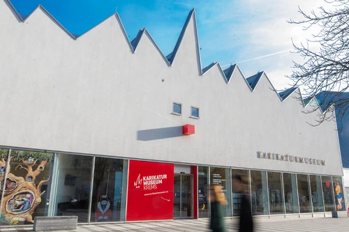 Moderne Museumsfassade mit gezacktem Dach und großem roten Eingangselement, davor Passanten.