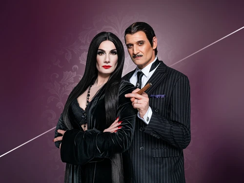 Zwei Personen posieren als Morticia und Gomez Addams in klassischen Kostümen vor einem dunkellila Hintergrund mit floralem Muster.
