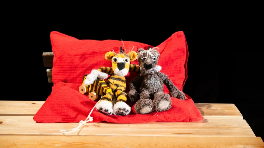 Zwei weiche Tierpuppen – ein Tiger mit Tigerente an der Hand und ein Bär – sitzen eng nebeneinander auf einem roten Kissen auf einem Holztisch vor schwarzem Hintergrund.
