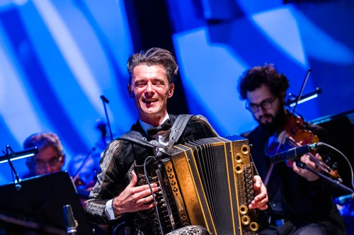 Konzertfoto von Herbert Pixner mit einer Ziehharmonika, hinter ihm zu sehen: das Tonkünstler Orchester Niederösterreich auf einer blau beleuchteten Bühne.