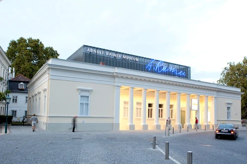 Frontansicht des Arnulf Rainer Museums mit klassizistischer Fassade und beleuchtetem Eingangsbereich, darüber moderne Neonaufschrift.