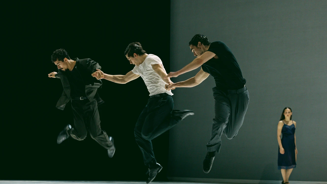 Szenenfoto von "Obsidian", einer Choreografie von Bobbi Jene Smith und Or Schraiber. Zu sehen sind Tänzer:innen des Ballet BC, drei von ihnen im Bildvordergrund, die gleichzeitig hochspringen, im Hintergrund steht eine Tänzerin im schwarzen Kleid vor dem grauen Bühnenhintergrund.