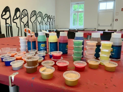 Tisch mit zahlreichen offenen Acrylfarben in Flaschen und kleinen Bechern, sortiert in verschiedenen bunten Farbtönen in einem Atelierraum.