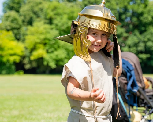 A small child is wearing a golden Roman helmet in front of green trees