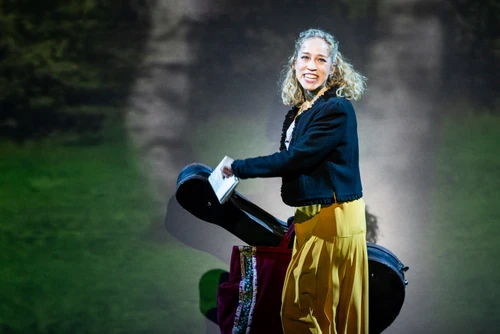 A woman in historical clothing with a light-coloured blouse, dark jacket and yellow skirt stands on a green meadow in front of blurred trees. She holds a guitar in front of her and looks slightly to the side, giving the scene a musical and nature-loving impression.