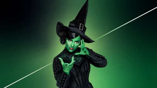 Grüner Hintergrund. Frau mit grün gefärbtem Gesicht und spitzem schwarzem Hut sowie schwarzem Kleid blickt in die Kamera, vor ihrem Gesicht ihre Hände gespreizt. Nahaufnahme.