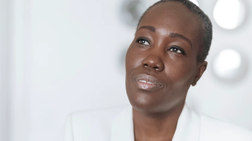 Chanda Rule sitzt mit weißem Blazer gekleidet, nachdenklich auf einem weißen Sofa, während sie mit ruhigem, leicht geheimnisvollem Blick vor hellem Hintergrund an der Kamera vorbeischaut.