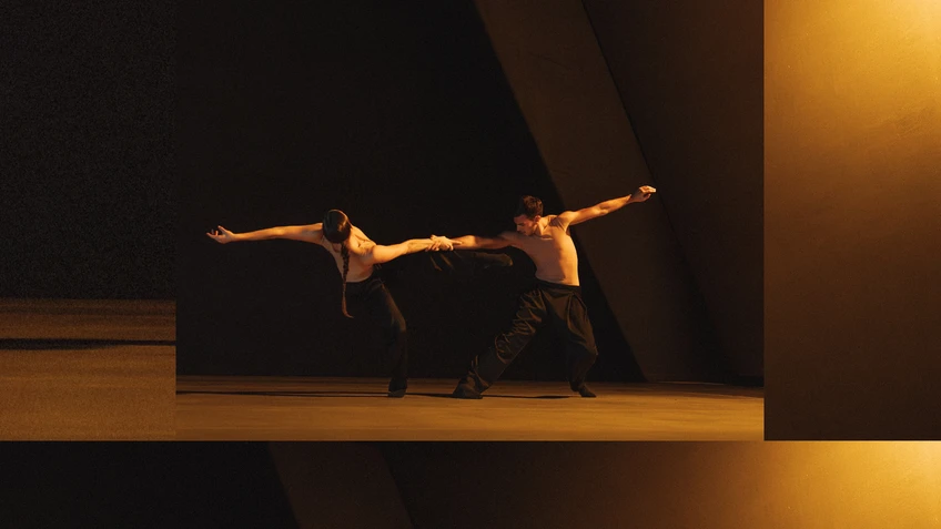 Performancefoto der Choreografie "SWAY" von Medhi Walerski. Zu sehen sind  zwei Tänzer:innen des Ballet BC, gekleidet in schwarzer Hose und weißem Shirt, vor schwarzem Bühnenhintergrund, in einem raumgreifenden Tanzschritt, beleuchtet mit gelbem Bühnenlicht. 