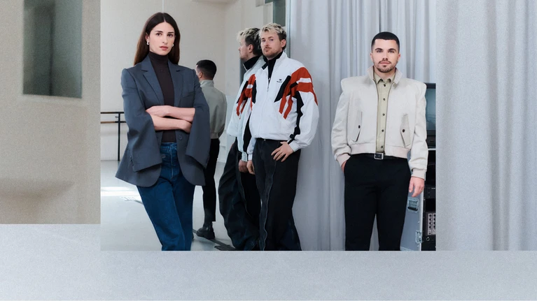 Foto des Kollektivs "(LA)HORDE". Die drei Künstler:innen stehen in einem Proberaum, hinter ihnen ist ein hellgrauer Vorhang und ein großer Spiegel zu sehen.