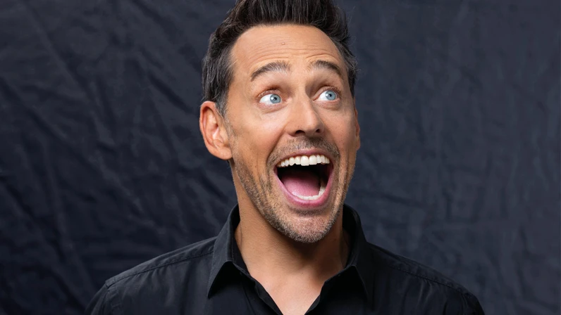 Tricky Niki steht mit schwarzem Hemd vor dunklem Hintergrund und lacht mit weit geöffnetem Mund nach rechts oben, als wäre er überrascht oder begeistert.