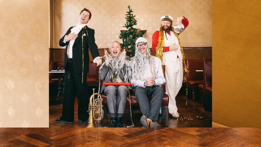 A photo of the musicians Sebastian Fuchsberger, Leonhard Paul, Michael Hornek and Thomas Gansch in an empty coffee house, with a Christmas tree behind them; the cheerful figures are partly draped in tinsel.