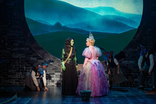 Green-skinned woman with black hair and black clothing and blonde woman in an exaggerated pink princess dress and huge tiara, facing each other as if in conversation, masked figures in the shadows, projections of a misty hilly landscape in the background