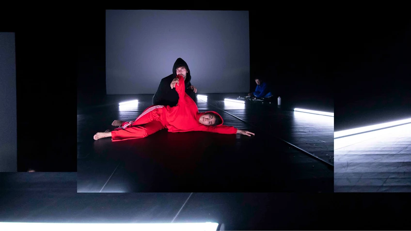 Performancefoto von "Rotkäppchen", choreografiert von Sylvain Huc. Zu sehen sind zwei Tänzer:innen, eine:r davon in einem knallroten Sportanzug, der:die andere in einem schwarzen Sportanzug.