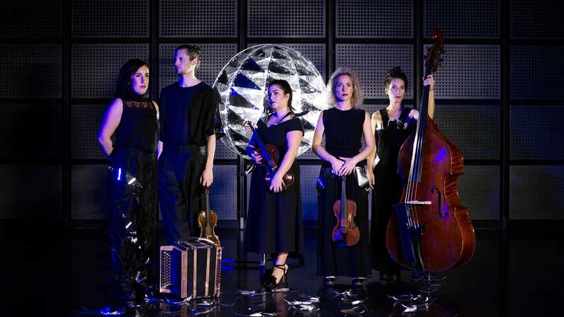Die fünf Musiker:innen von ALMA stehen in eleganten schwarzen Outfits vor einer metallisch glänzenden, kuppelförmigen Lichtinstallation, halten ihre Instrumente – Harmonium, Violinen, Bratsche und Kontrabass – und blicken ernst in die Kamera.