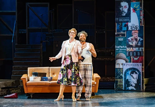 Two people are standing in front of a brown sofa on a theatre stage. One is wearing a light-coloured top and a patterned skirt; the other is wearing a checked garment. Both are at the centre of the scene, standing in front of a backdrop featuring posters, stairs and scaffolding. The stage is bathed in blue light, whilst the sofa and the figures are highlighted by brighter lighting. The composition focuses attention on the encounter between the two people.
