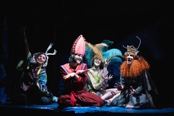 Szenenbild aus "Don Quijote". Vier Schauspieler:innen in bunten Kostümen, mit Perücken und Kopfschmuck werden von einem Lichtstrahl erleuchtet.