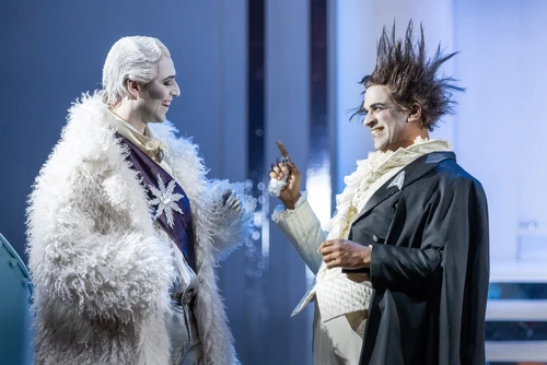 Two figures stand facing each other on a brightly lit stage. The figure on the left wears a long, white, fur-like costume with a decorative star-shaped element on the chest and a light-coloured, elegant undergarment. The figure on the right wears a dark cape over a light-coloured costume with a ruffled collar and holds one hand slightly raised, as if emphasising a point. Both stand in front of a bright stage backdrop with geometric structures, bathed in cool blue and white light.