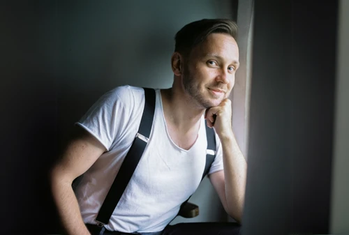 Mann in weißem T-Shirt und Hosenträgern sitzt an einem Fenster und stützt sich auf seine linke Hand